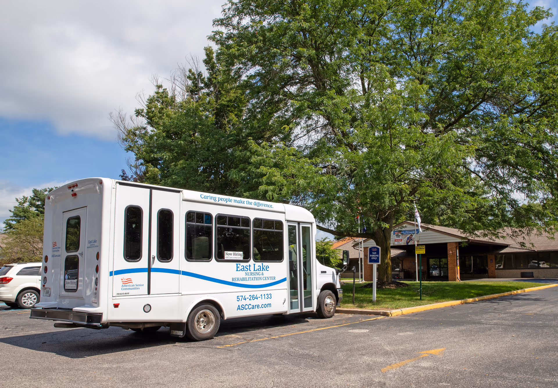 A white shuttle bus parked in front of a single-story building with a covered entrance. The bus has signage for East Lake Nursing & Rehabilitation Center and contact information. There are trees and a partly cloudy sky in the background.