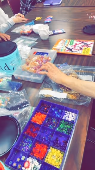 Several people sitting around a table engaged in a bead crafting activity. The table is covered with various containers and bags of colorful beads, a pair of scissors, a magazine, and some small white bowls.