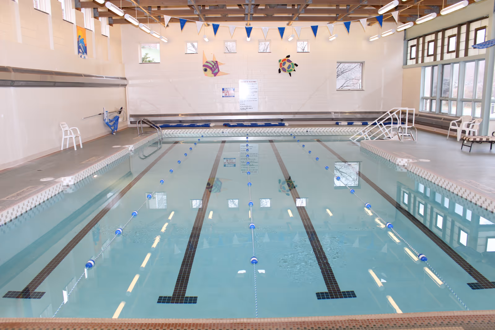 Indoor swimming pool with clear water and lane dividers, surrounded by a tiled deck with white plastic chairs and benches. The room has large windows on the right side, a high ceiling with wooden beams, and colorful fish decorations on the far wall.