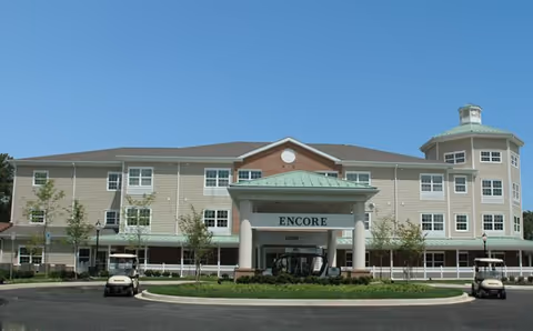 Front exterior view of a large senior living facility named Encore with a covered entrance, multiple windows, and a round tower-like structure on the right side. There are two golf carts parked in front and some small trees and landscaping around the building.