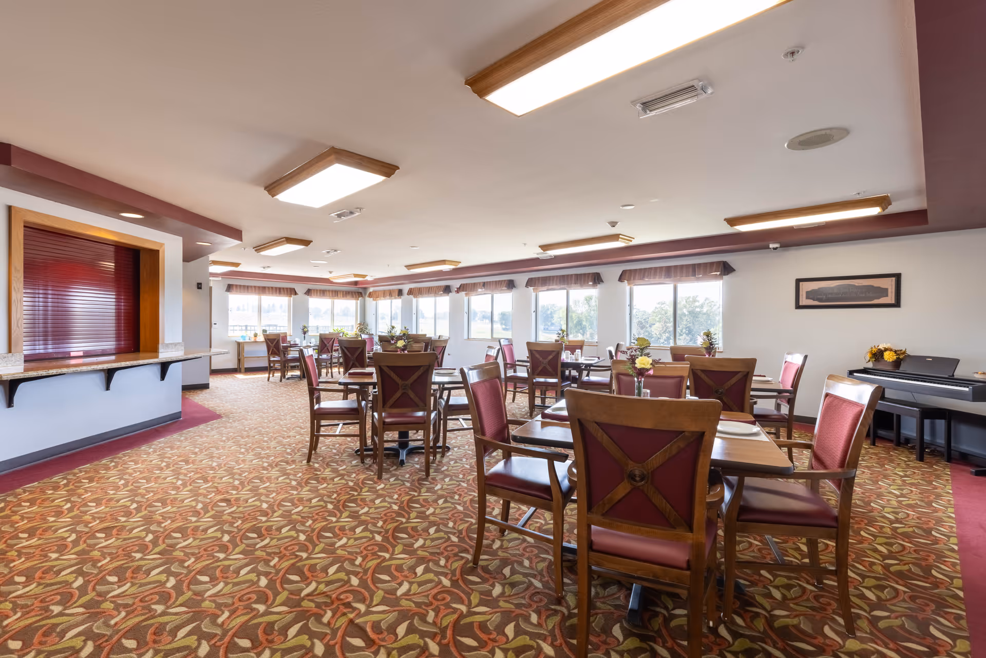 A spacious dining room with multiple wooden tables and chairs featuring red cushioned seats. The room has large windows letting in natural light, floral arrangements on the tables, patterned carpet flooring, and ceiling lights. There is a piano against the wall on the right and a serving counter with a closed red shutter on the left.