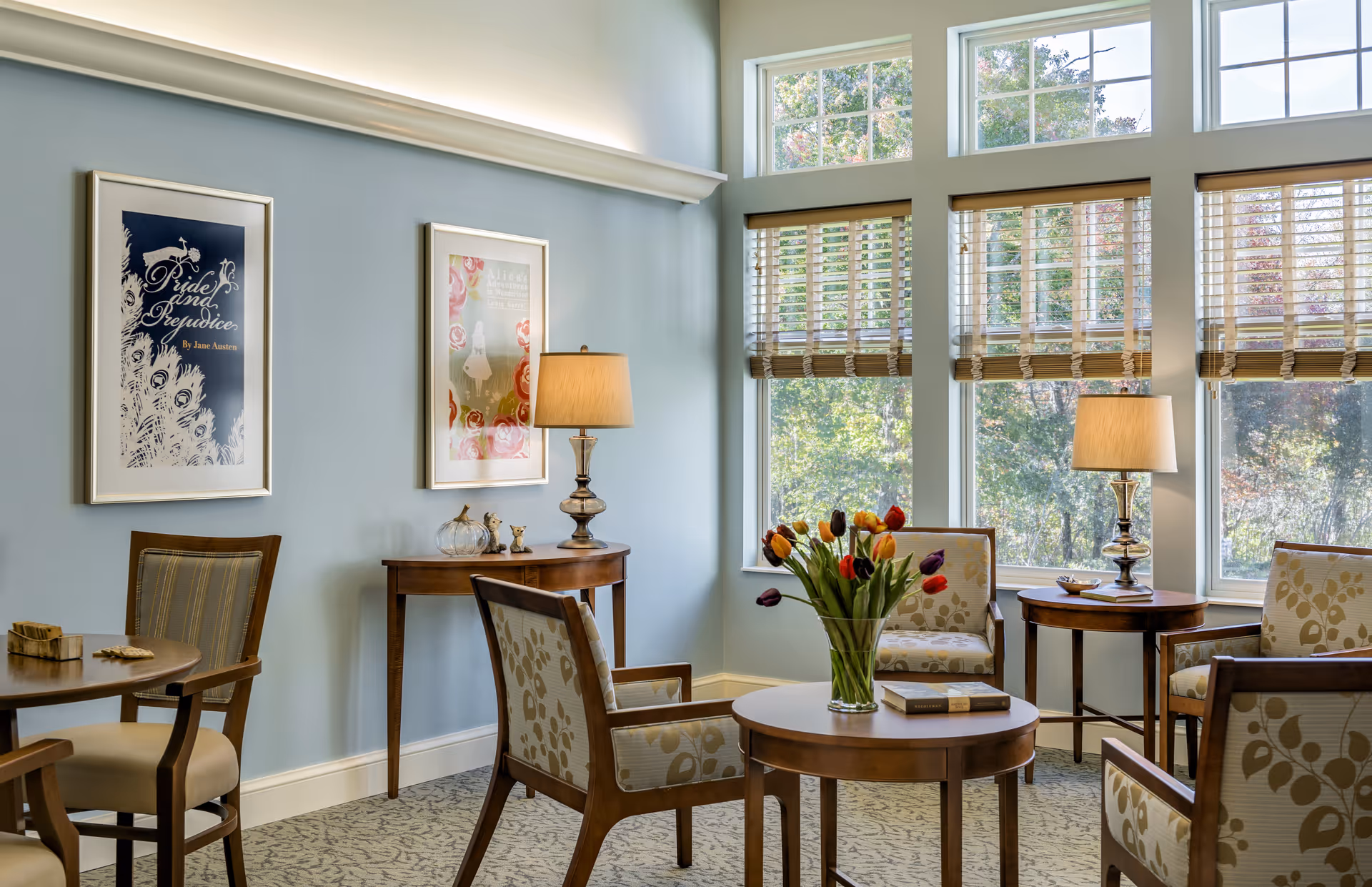 A cozy sitting area with wooden chairs upholstered in floral fabric arranged around a round wooden table with a vase of colorful tulips. The room has large windows with wooden blinds letting in natural light, two framed artworks on a light blue wall, and two table lamps on small wooden side tables.