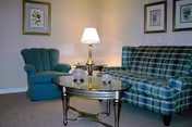 A cozy living room with a blue and green plaid sofa, a teal upholstered armchair, a glass-top coffee table with decorative items, a brass table lamp on a side table, and framed artwork on the beige walls.