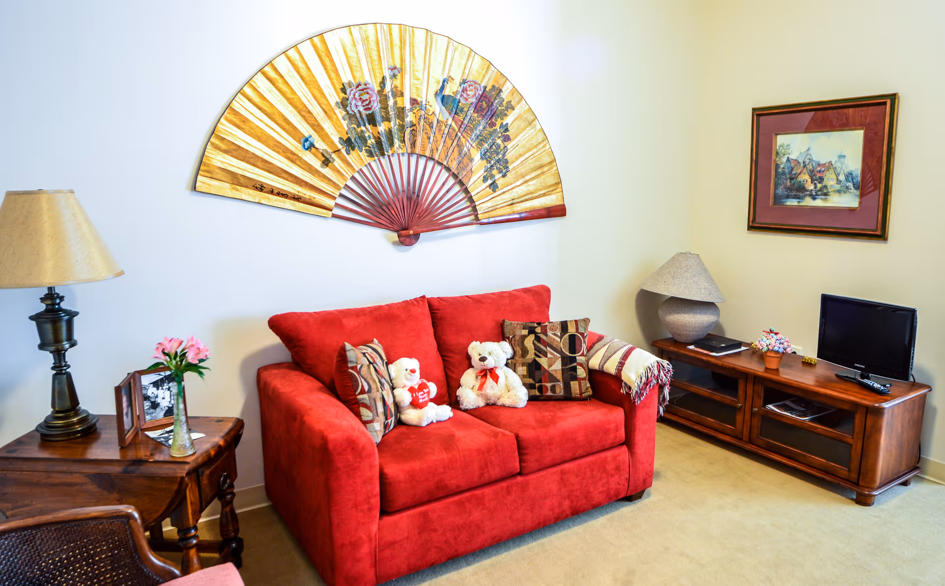 A cozy living room with a bright red loveseat adorned with patterned pillows and two teddy bears. Above the loveseat is a large decorative fan with floral designs. To the left is a wooden side table with a lamp, a vase with pink flowers, and framed photos. To the right is a wooden TV stand with a small flat-screen TV, a lamp, a potted plant, and a framed picture hanging on the wall above.