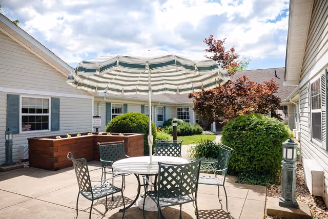 Outdoor patio area at Arden Courts - ProMedica Memory Care Community (Silver Spring) featuring a round table with a striped umbrella and four metal chairs. Surrounding the patio are bushes, a tree with red leaves, and raised garden beds. The building exterior is light-colored with windows and shutters under a partly cloudy sky.