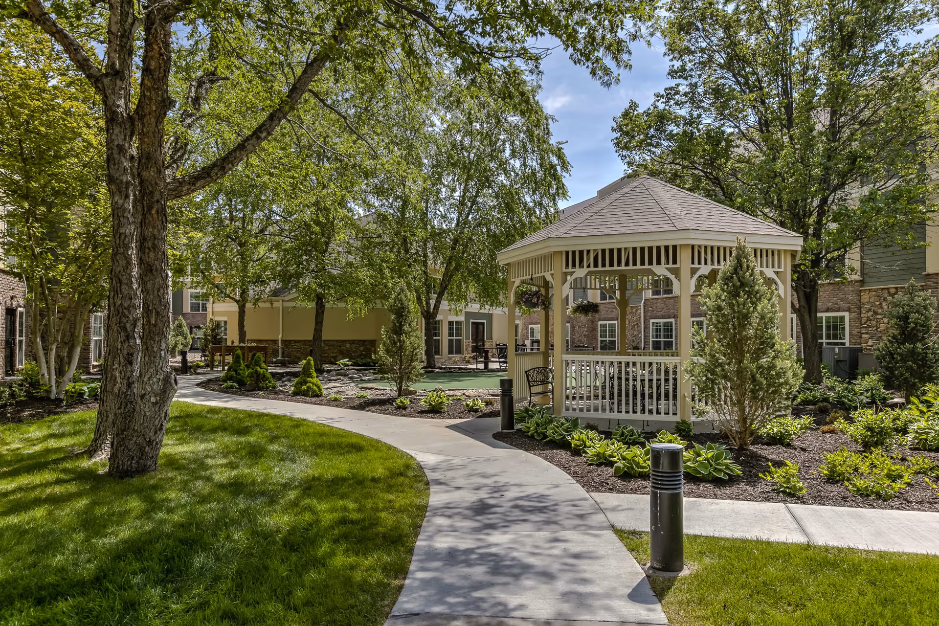 A peaceful outdoor garden area at Remington Heights Retirement Community featuring a curved concrete pathway, green grass, trees, shrubs, and a white wooden gazebo with benches inside. The surrounding buildings have stone and siding exteriors with multiple windows.