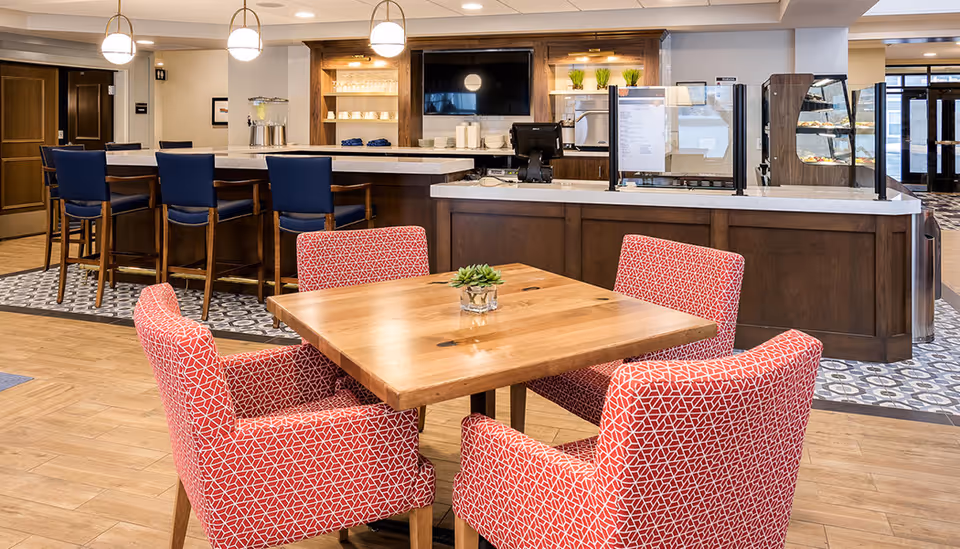 Wood table with four red patterned upholstered chairs in a dining/lounge area facing a service counter with bar stools and a display case.