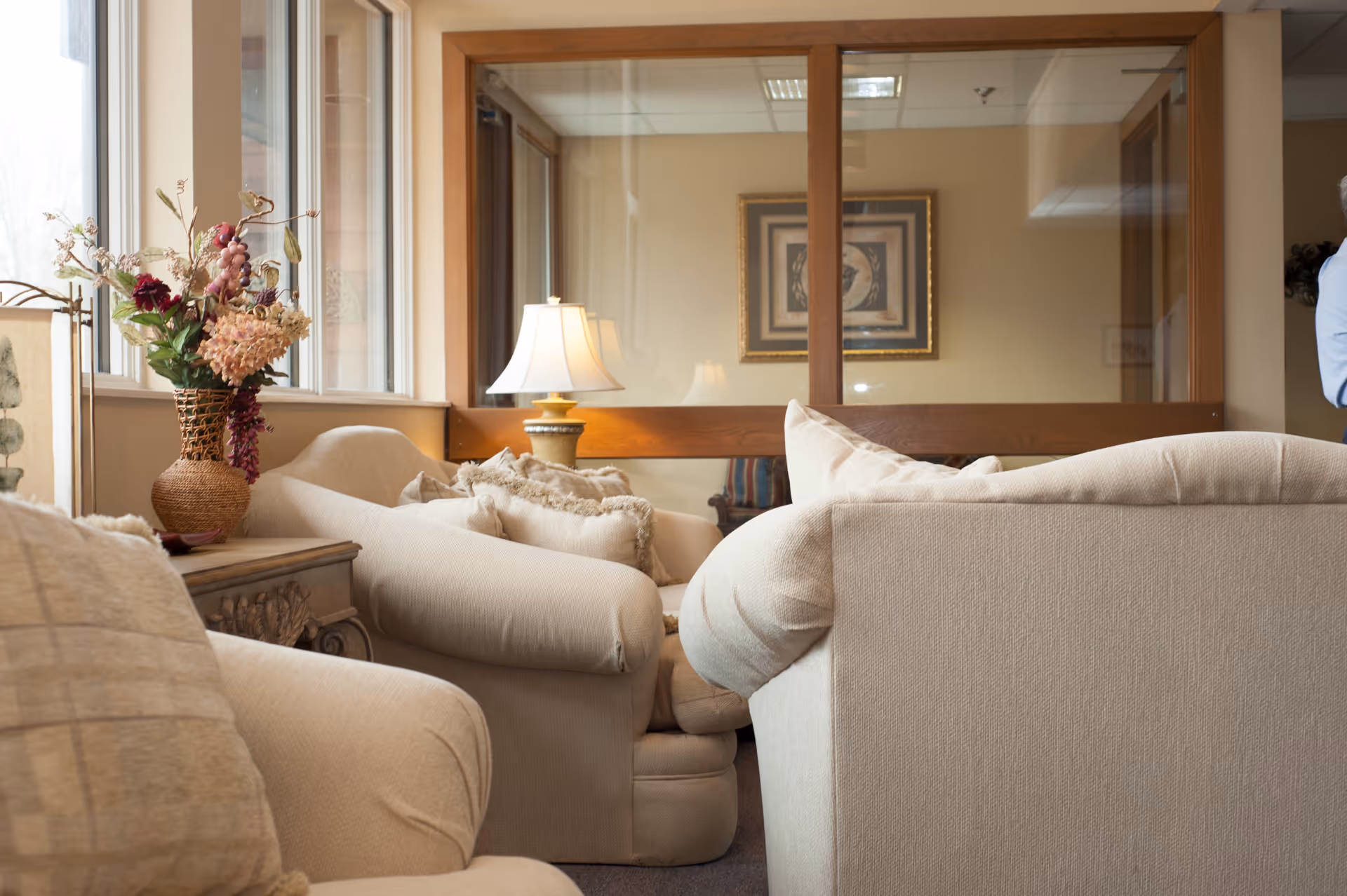 A cozy living room area with beige upholstered armchairs and sofas arranged around a wooden side table with a decorative vase of flowers. A table lamp with a white shade is placed on the side table. The room has large windows letting in natural light and a framed artwork on the wall behind glass panels.