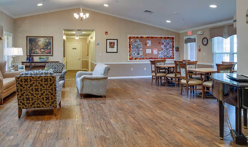 A spacious senior living common area with wooden flooring, several upholstered armchairs, a sofa, a wooden dining table with chairs, a bulletin board with photos and notes on the wall, a piano on the right side, and large windows letting in natural light.