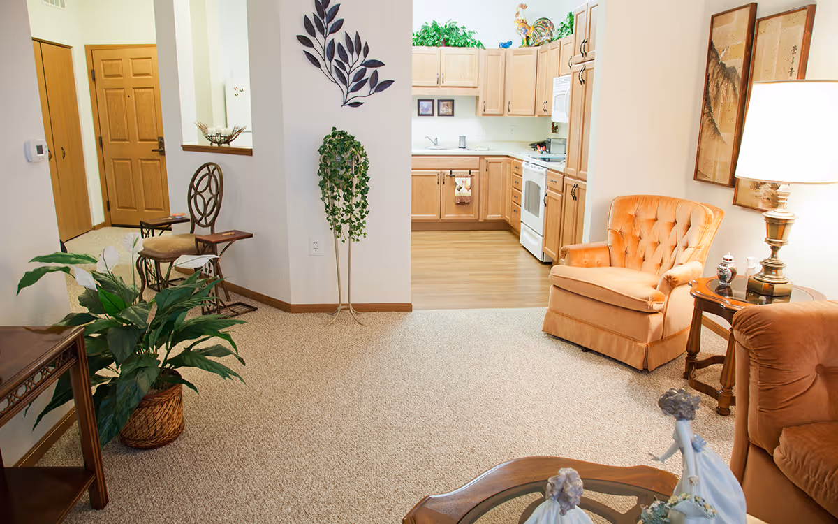 A cozy senior living interior featuring a small kitchen with wooden cabinets and white appliances, an adjacent living area with an orange upholstered armchair, a side table with a lamp, decorative wall art, a potted plant, and a chair with a small table near the entrance door.