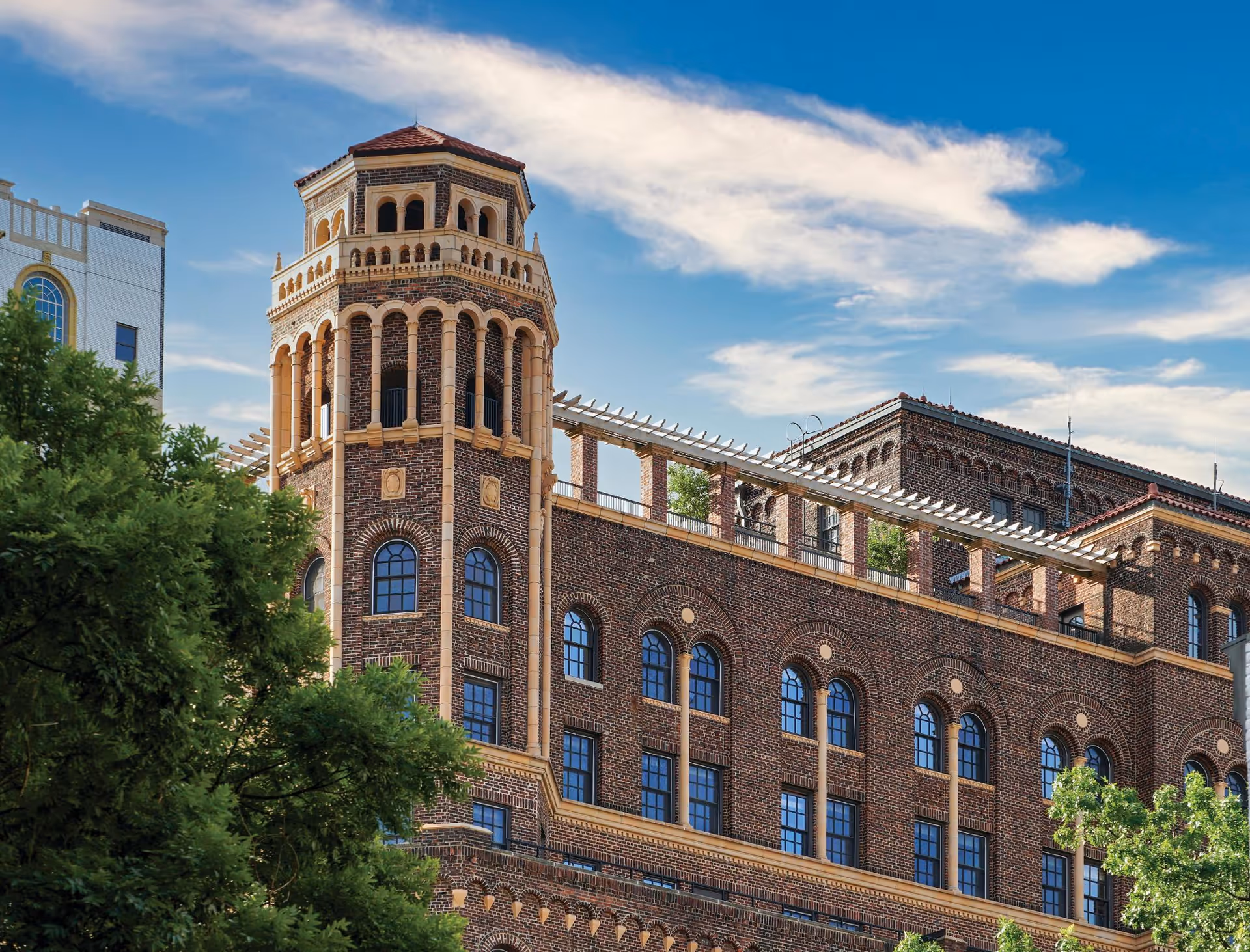 Exterior view of a multi-story brick building with arched windows and a decorative tower under a partly cloudy blue sky, with green trees in the foreground.