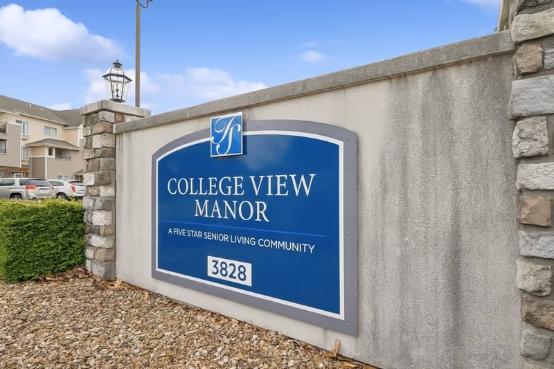 Entrance sign for College View Manor, a five star senior living community, mounted on a stone and concrete wall with a lamp post on top. In the background, there are parked cars and part of the building visible under a blue sky with some clouds.