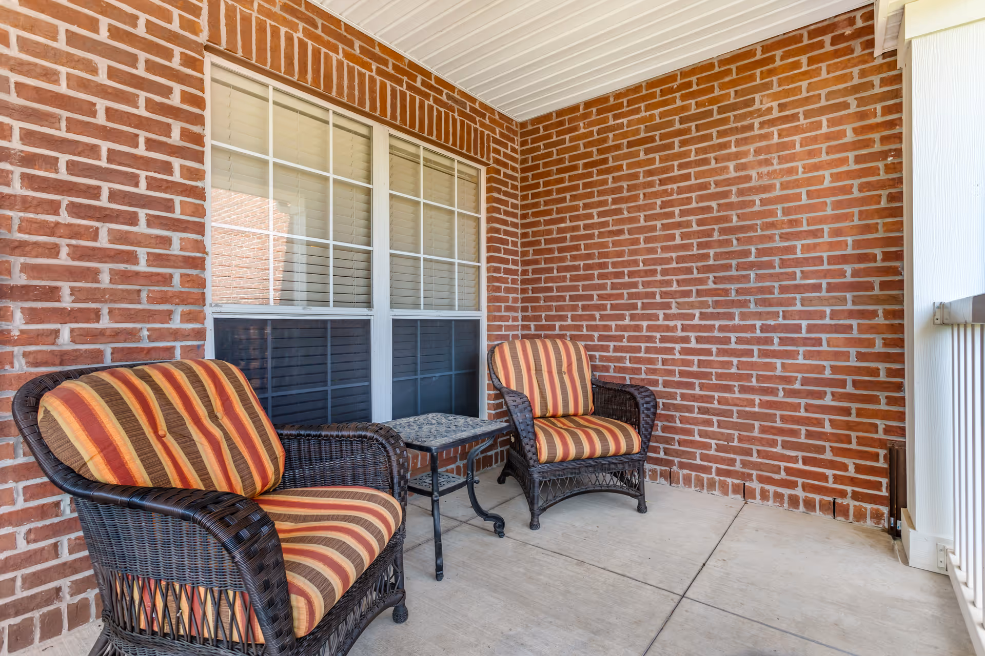 Two wicker chairs with striped cushions and a small table on a covered brick porch outside a window.