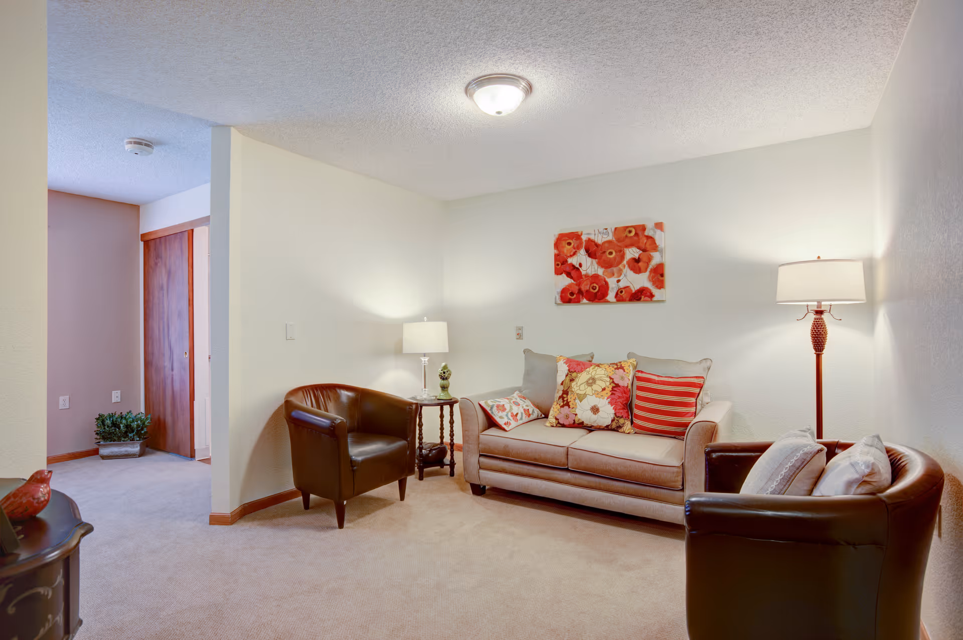 Well-lit living room with a beige sofa, two leather armchairs, floral pillows and artwork, and standing and table lamps.