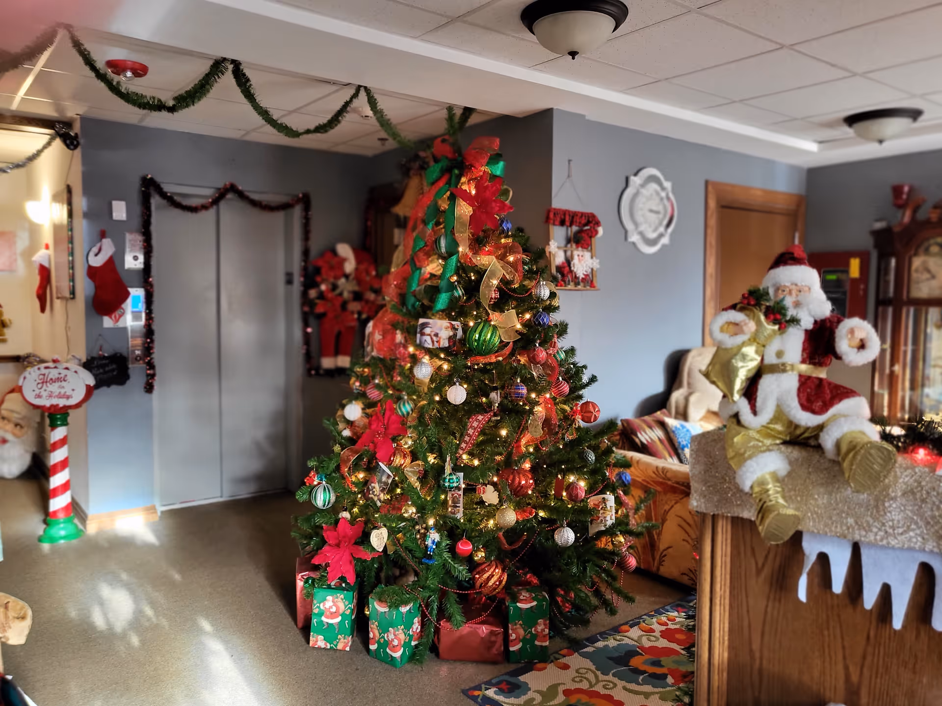 A holiday-decorated interior lobby with a large Christmas tree, wrapped gifts, a Santa figure, and elevator doors in the background.