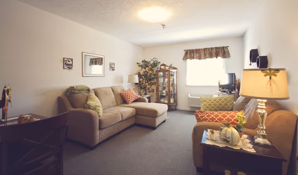 A cozy living room with beige couches adorned with patterned pillows, a wooden side table with a lamp and decorative items, a glass display cabinet with collectibles, a window with floral valance, and a carpeted floor.