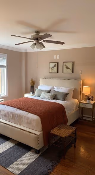A cozy bedroom with a large bed featuring a white upholstered headboard and white bedding topped with a rust-colored blanket and several pillows. There are two framed bird pictures hanging above the bed. A ceiling fan with lights is mounted on the ceiling. Next to the bed is a nightstand with a lamp and a small plant. A woven stool is placed at the foot of the bed, and a striped rug lies on the wooden floor.