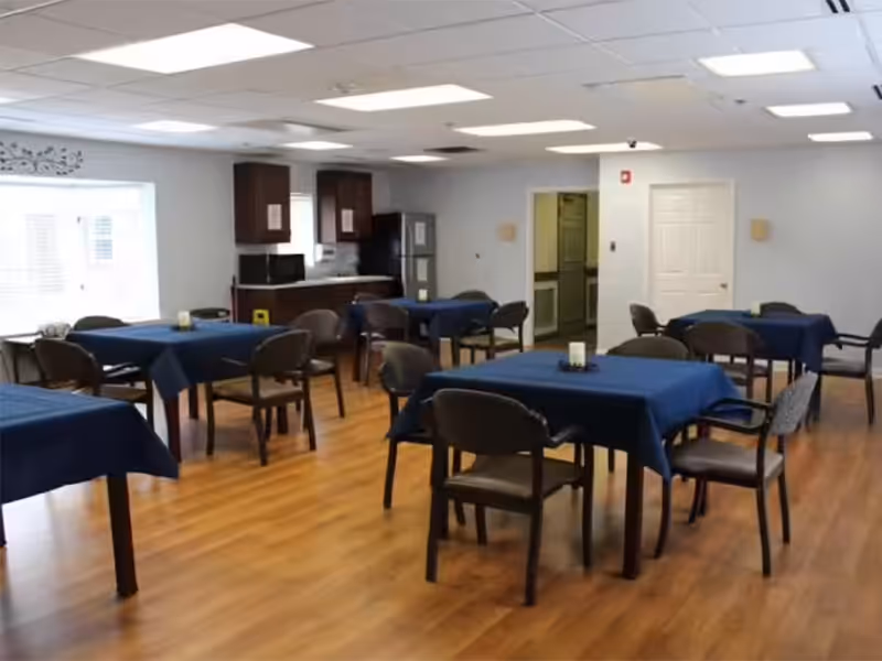 A dining room with multiple square tables covered in dark blue tablecloths, each surrounded by four chairs. The room has wooden flooring, white walls, and a ceiling with recessed lighting. There is a kitchenette area with cabinets, a microwave, and a refrigerator in the background, and a window letting in natural light on the left side.