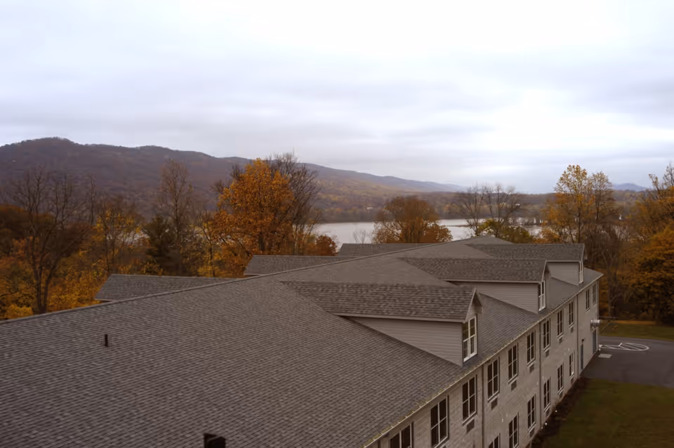 Aerial view of a large senior living facility building with a gray shingled roof, surrounded by autumn trees with colorful foliage. In the background, there are hills and a body of water under a cloudy sky.