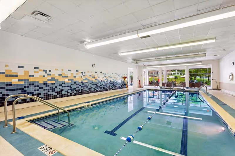 Indoor swimming pool with lane divider, tile mosaic wall, handrails, and seating visible through large windows.