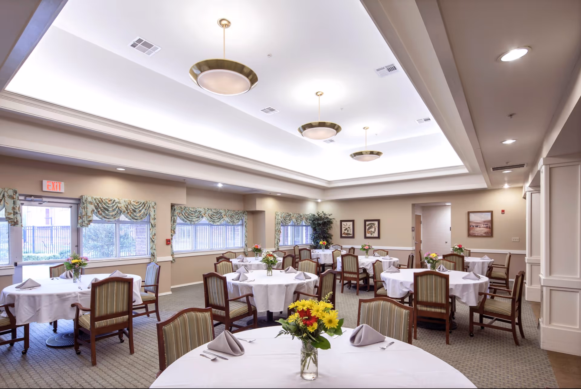 Bright dining room with round tables set with white tablecloths, chairs, and floral centerpieces under recessed lighting.