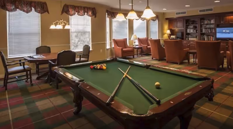 A communal game room with a pool table in the foreground and seating, tables, and bookshelves in the background.
