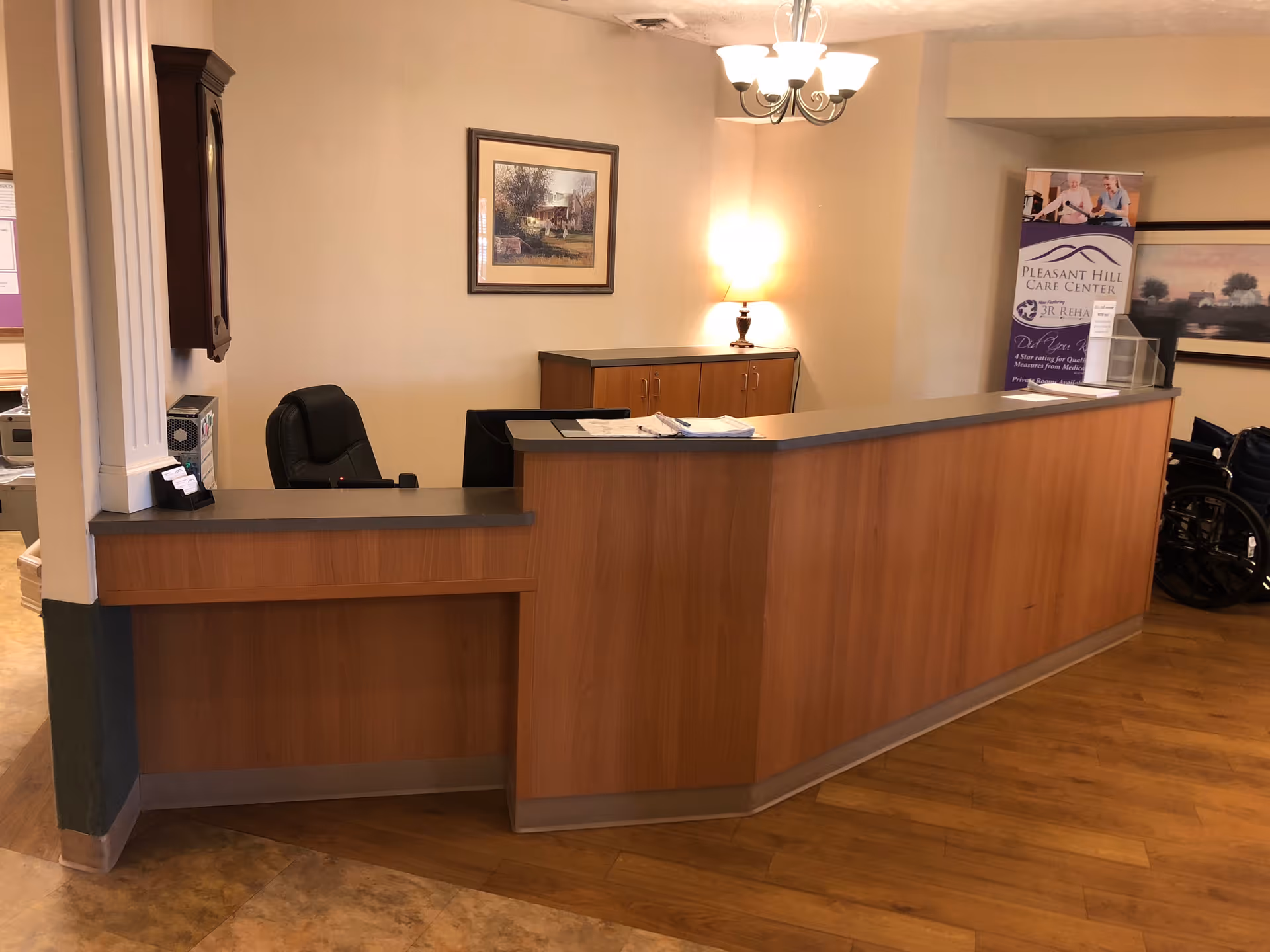 Reception area with a wooden front desk, a black office chair behind the desk, a table lamp on a cabinet against the wall, framed artwork on the wall, a chandelier light fixture on the ceiling, and a banner for Pleasant Hill Care Center in the background.