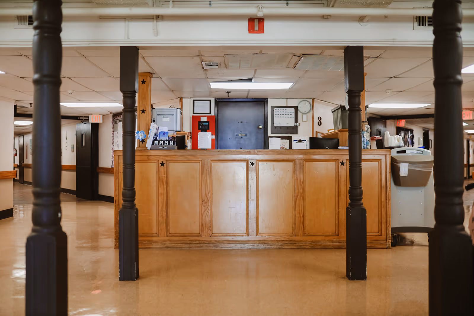 Reception desk area in a healthcare facility with wooden paneling and black pillars in the foreground. The desk has office supplies, a computer, and a trash bin nearby. Hallways extend on both sides with exit signs visible.