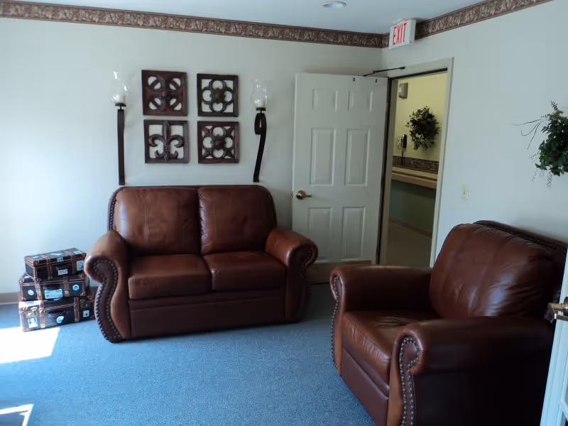Small sitting room with brown leather loveseat and armchair, wall decor, an open door and blue carpet.