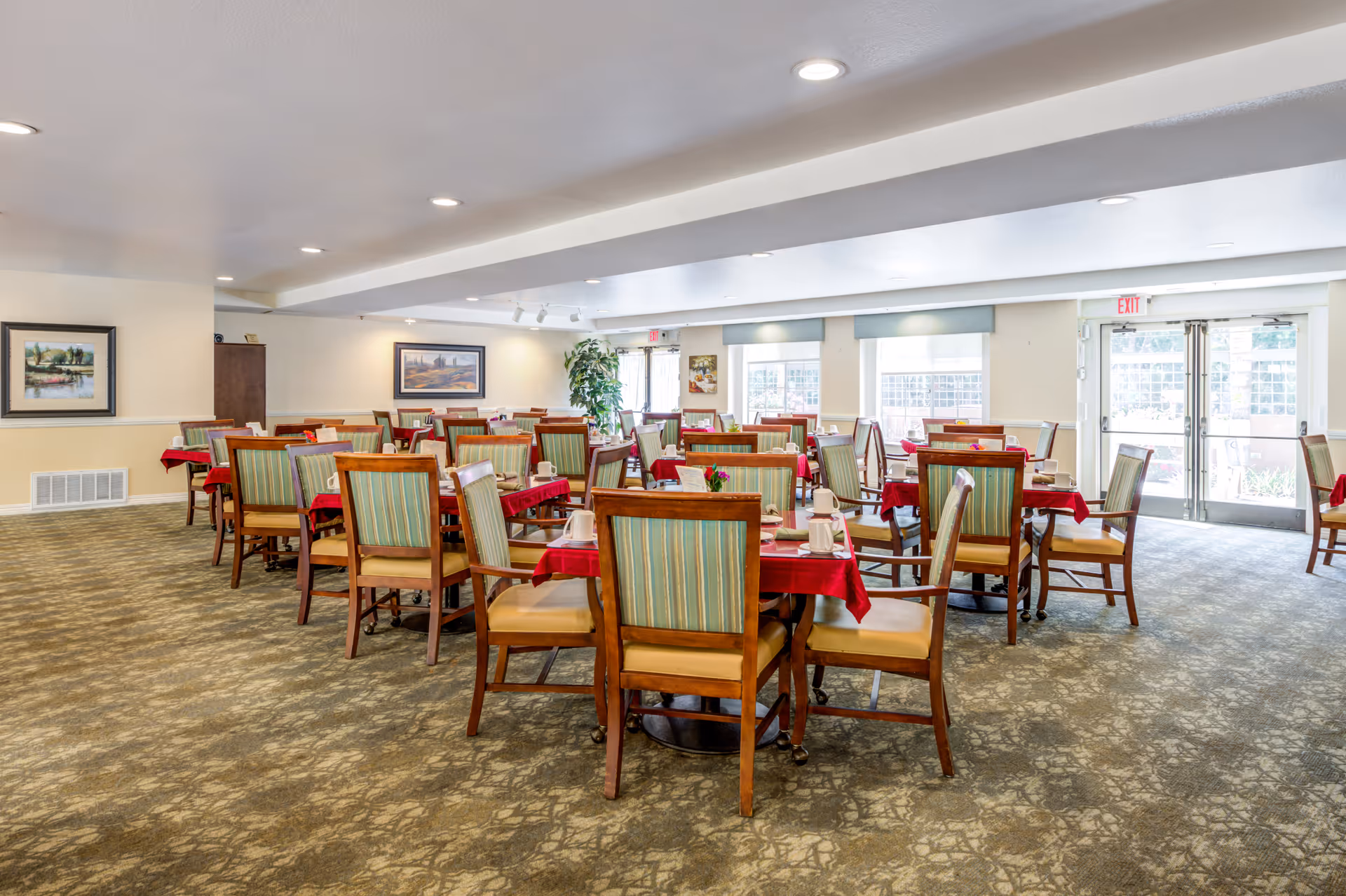 A spacious dining room in a senior living facility with multiple wooden tables covered with red tablecloths and surrounded by wooden chairs with green striped cushions. The room has large windows letting in natural light, framed paintings on the walls, and a carpeted floor with a patterned design.