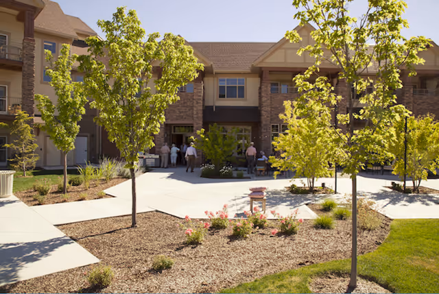 Outdoor courtyard area of a senior living facility with young trees, landscaped garden beds, and a paved walkway. Several people are gathered near the entrance of a two-story building with brick and beige siding under a clear blue sky.