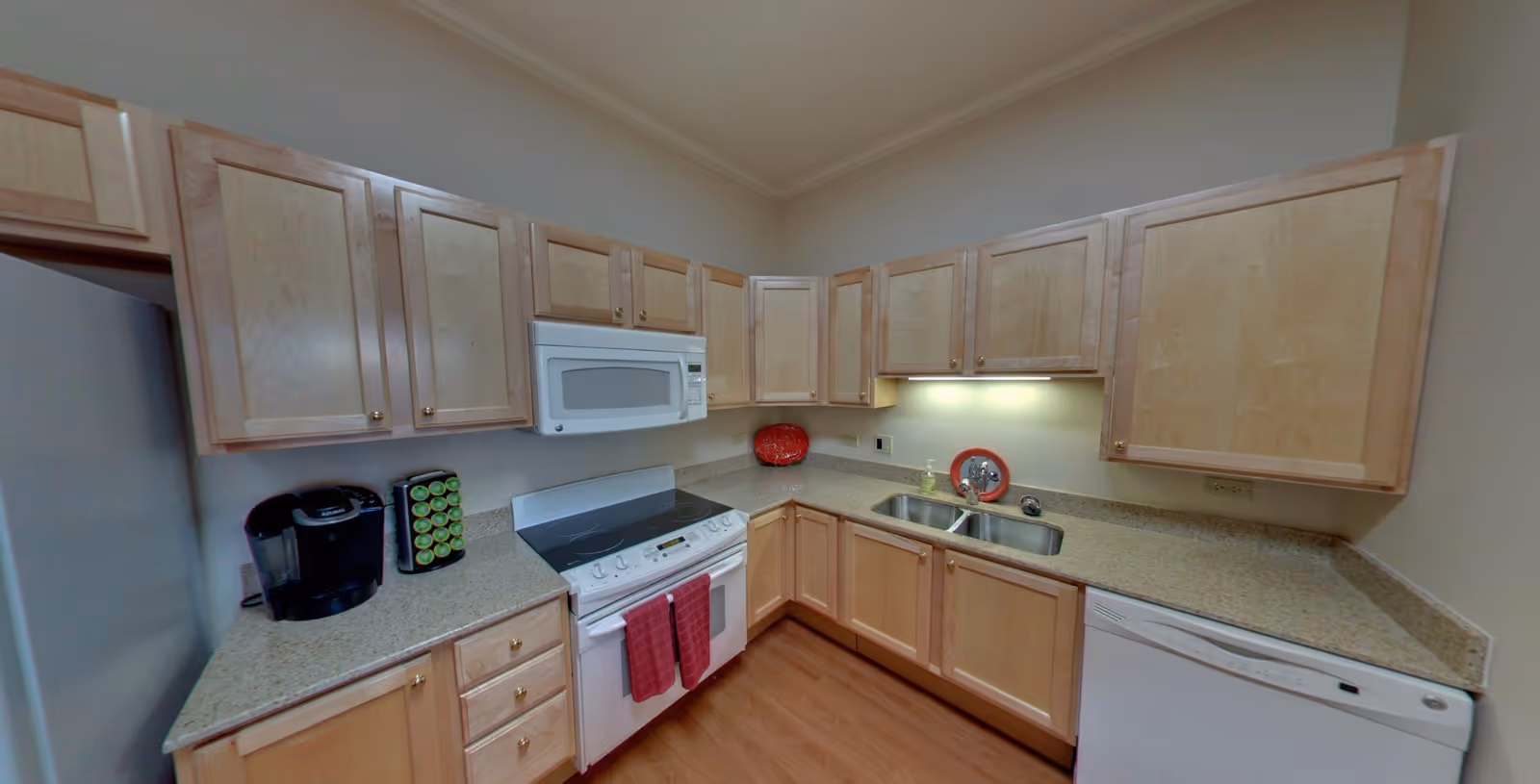 A kitchen with light wood cabinets, a white electric stove with a microwave above it, a double sink, a dishwasher, and a coffee maker on the countertop. The countertops are a speckled beige color and the floor is wood. There are two red towels hanging on the oven door and a red plate on the counter near the sink.