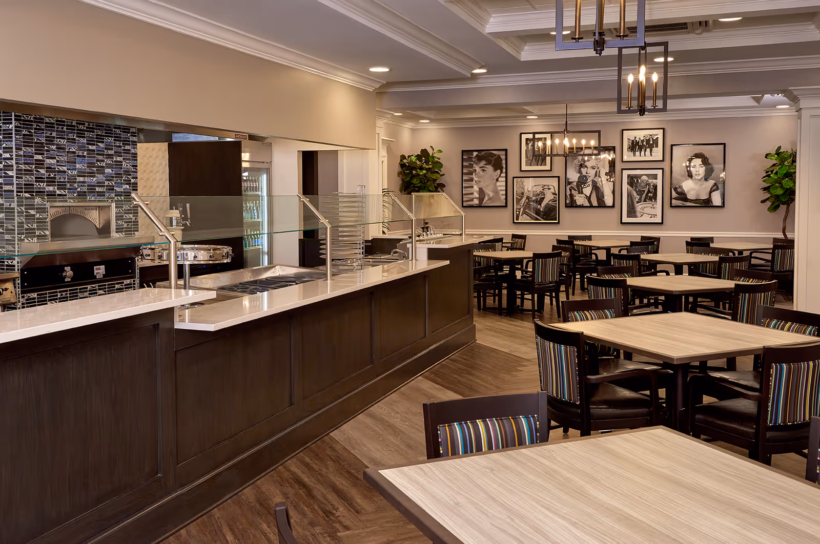 A dining area in an assisted living facility featuring multiple square wooden tables with dark wooden chairs that have striped cushions. There is a long serving counter with glass sneeze guards and a tiled backsplash behind it. The walls are decorated with black and white framed photographs of classic movie stars. The room has warm lighting with chandeliers and recessed ceiling lights, and there are potted plants in the corners.