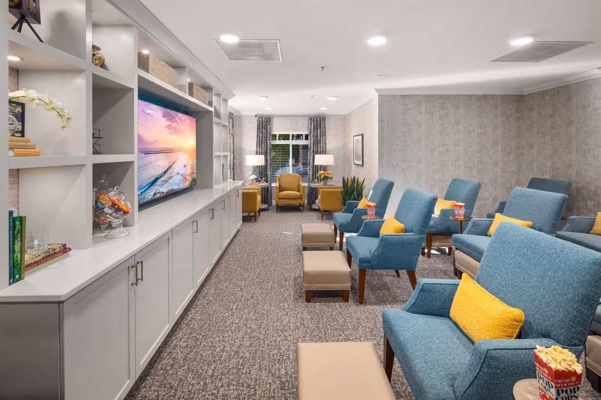 A cozy living room area in a senior living facility with multiple blue armchairs and matching ottomans, each with a yellow pillow. There are small side tables with popcorn containers on them. The room features a large built-in white shelving unit with a flat-screen TV displaying a sunset over the ocean. The far end of the room has two yellow armchairs near a window with patterned curtains and two table lamps.