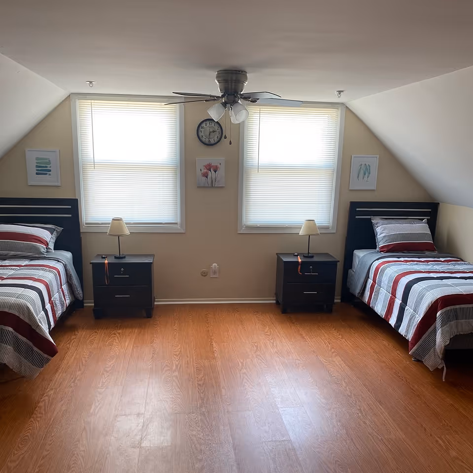 A bedroom with two single beds placed against opposite walls, each with striped bedding in shades of red, gray, and white. Between the beds are two black nightstands, each with a small lamp. Two windows with blinds are centered on the back wall, with a clock and two framed pictures hanging between and beside them. The room has wooden flooring and a ceiling fan with lights.