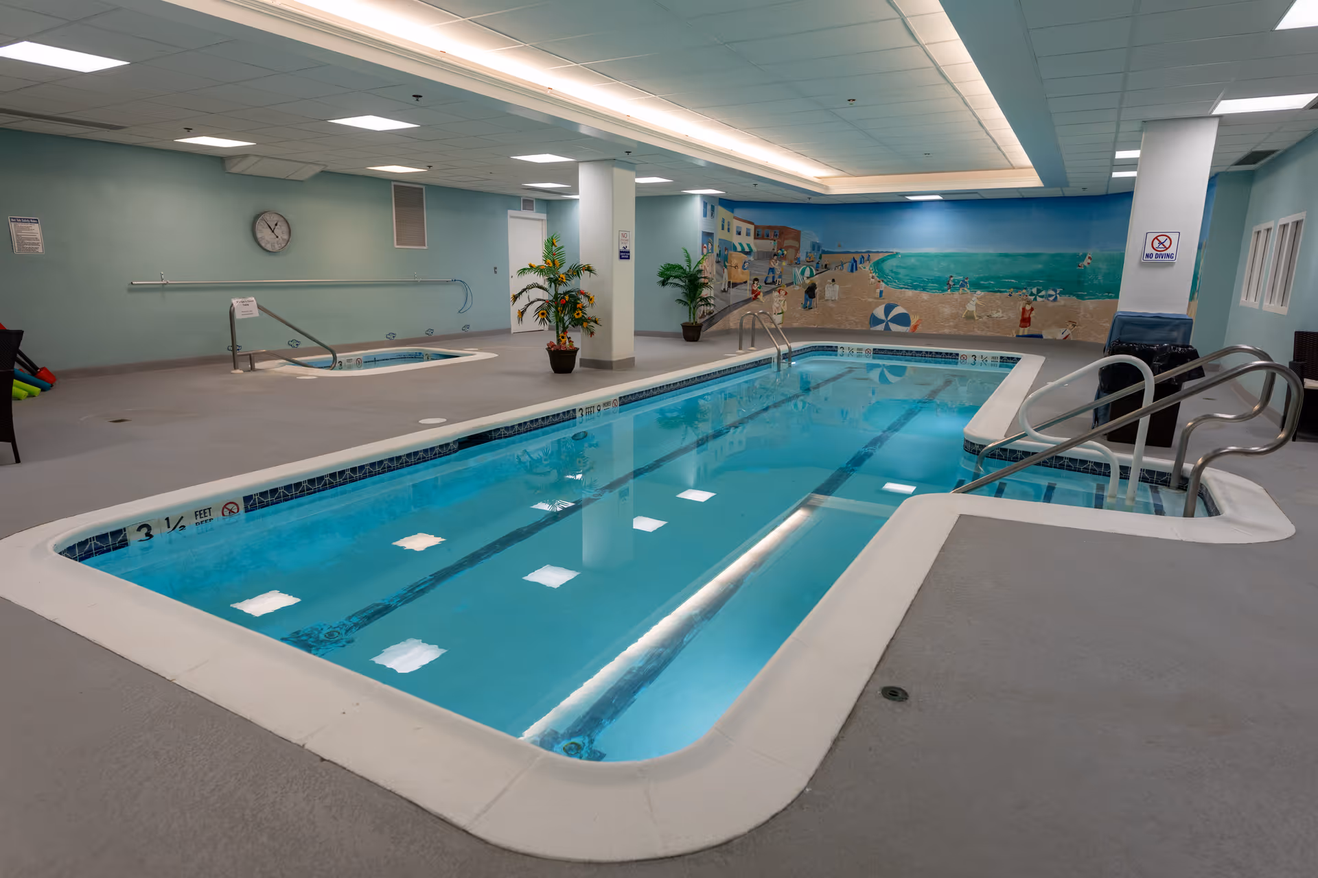 Indoor swimming pool area with a rectangular pool and a smaller hot tub. The room has light blue walls, a clock on the wall, potted plants, and a mural depicting a beach scene. There are handrails for entering the pool and hot tub, and the ceiling has recessed lighting.