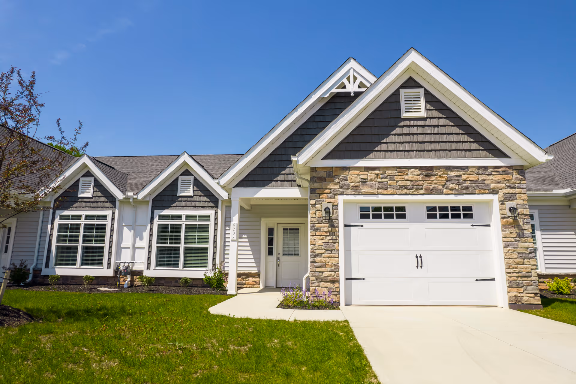 Exterior front view of a single-story residential building with a stone and siding facade, featuring a white garage door, multiple windows with white frames, a small covered entrance, and a well-maintained green lawn under a clear blue sky.