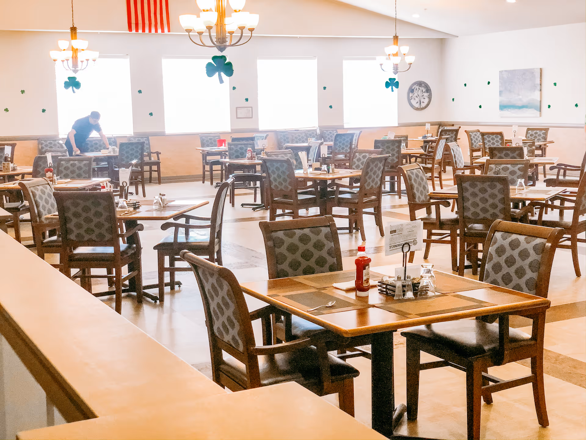 Open dining room in a senior living facility with many set tables and chairs, chandeliers, shamrock wall decorations, and a staff member setting a table.
