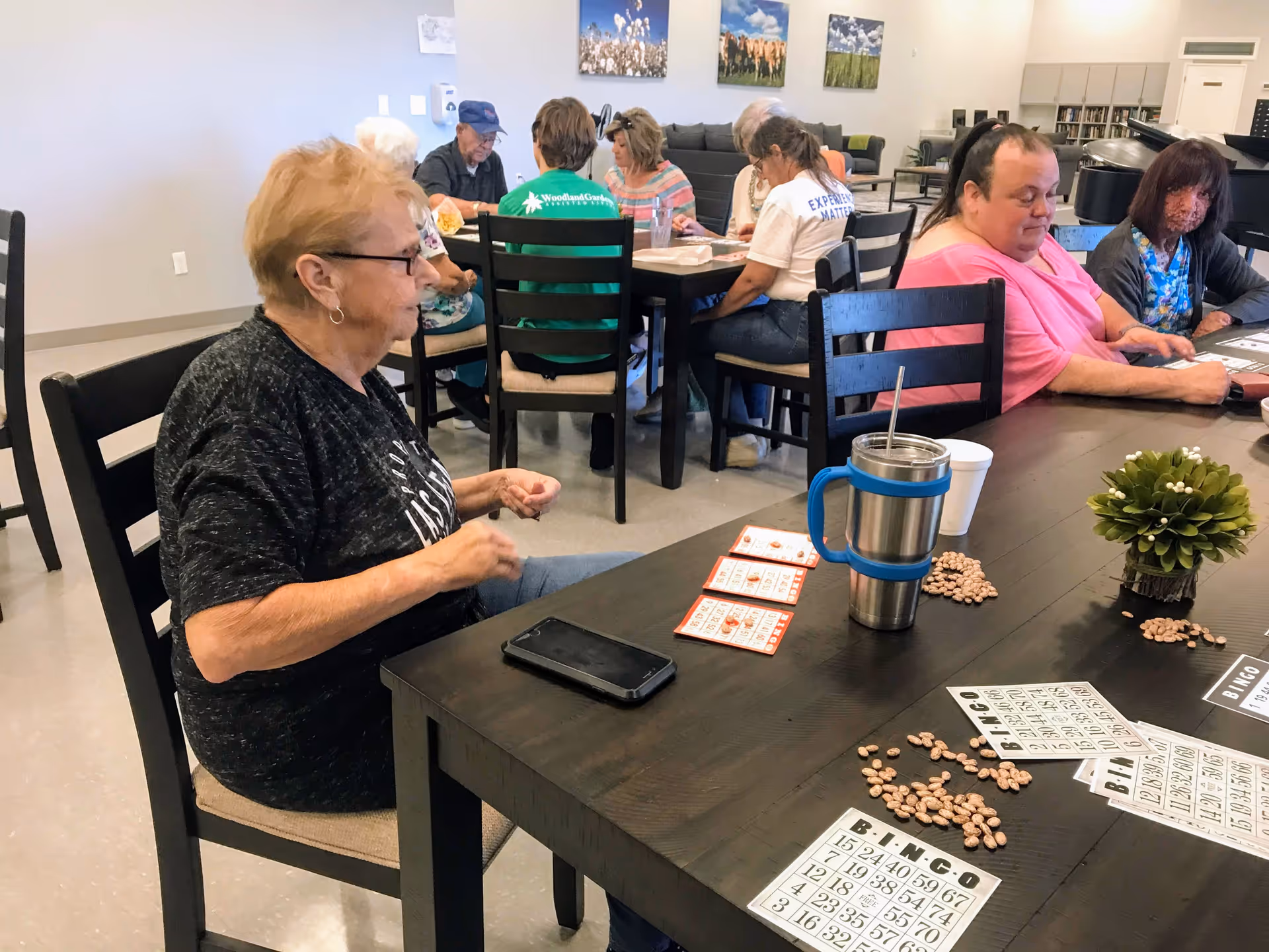 A group of elderly people sitting around tables in a common room playing bingo. Several bingo cards and beans are spread out on the tables. The room has simple decor with paintings on the wall and a small plant on the table.