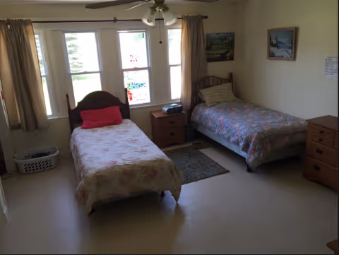 A simple bedroom with two single beds, each with floral bedspreads and pillows. Between the beds is a small wooden nightstand with a radio on top. The room has three windows with beige curtains, letting in natural light. There are two paintings on the wall and a small rug on the floor between the beds. A laundry basket is placed near the left bed.