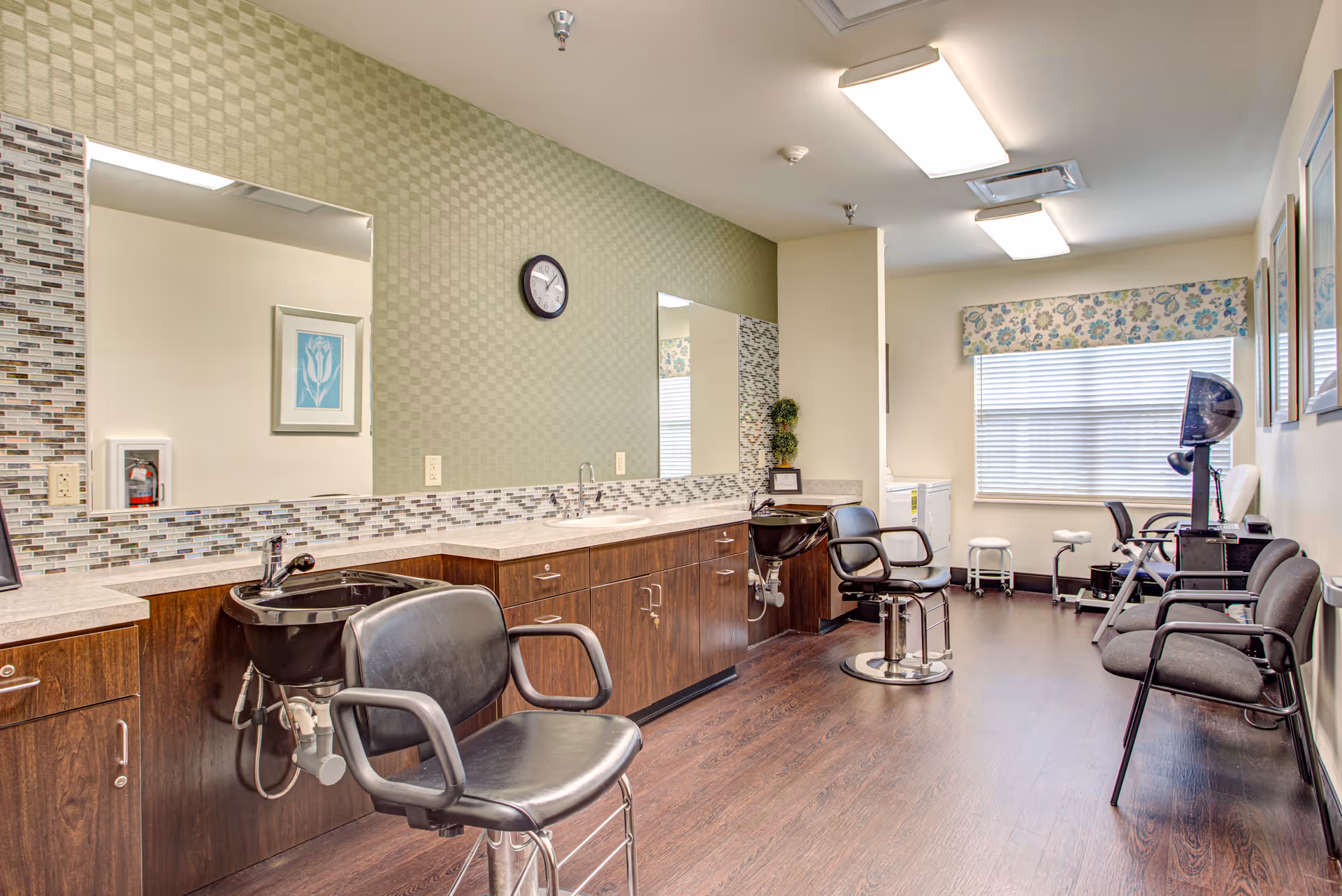 Interior of a hair salon area in a senior living facility with two black salon chairs in front of sinks and mirrors, wooden cabinetry, a clock on a green patterned wall, and additional seating along the opposite wall near a window with floral valance.