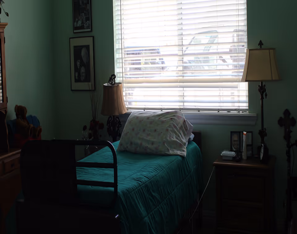 Single-occupancy bedroom with a bed with green quilt and rail, pillows, bedside tables with lamps, and a window with blinds.