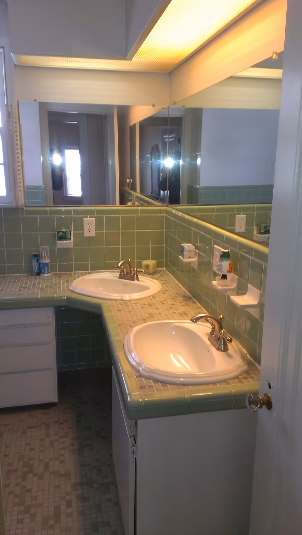 Bathroom with two sinks set in a green tiled countertop beneath a large mirrored wall and overhead lighting.