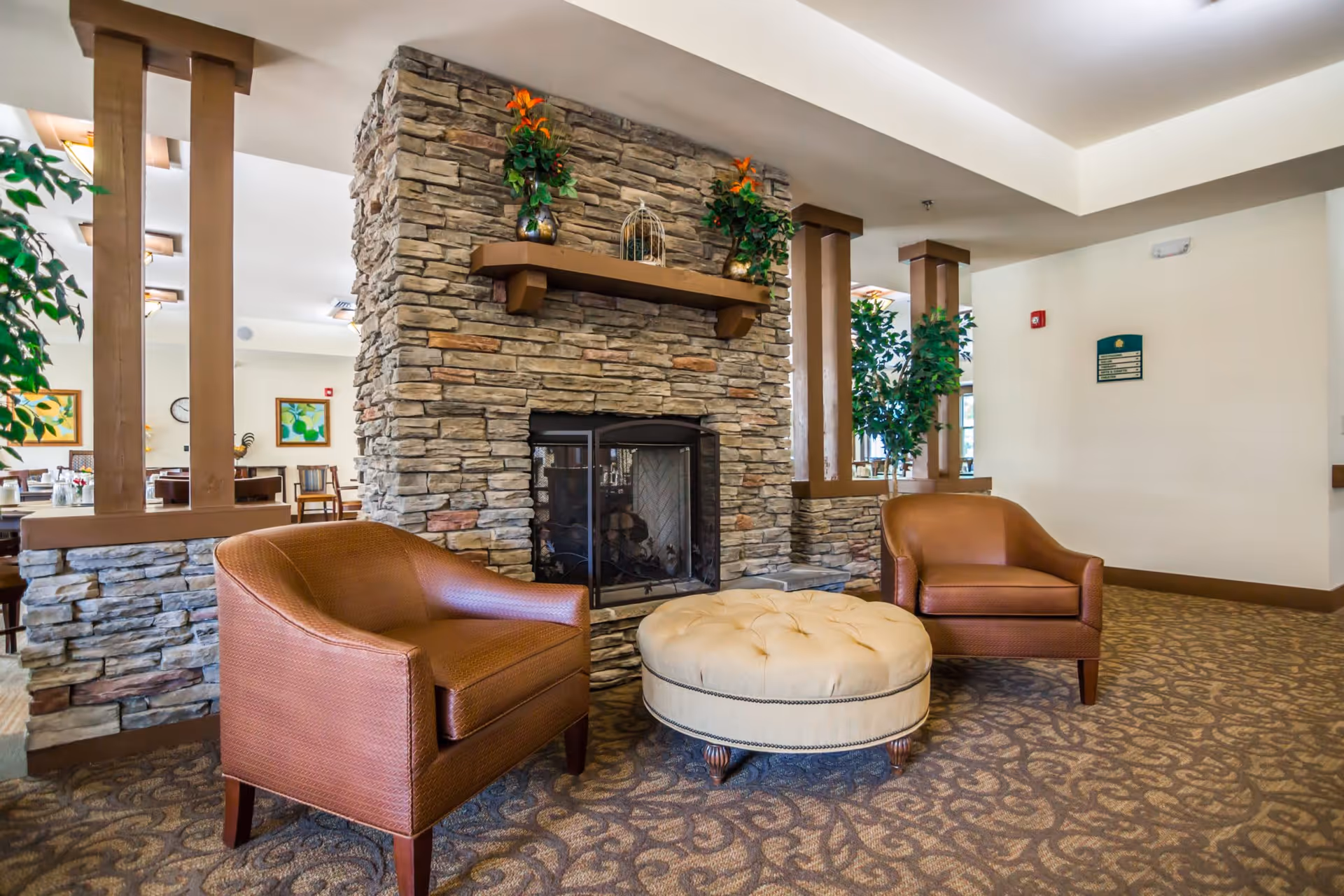 A cozy seating area in a senior living facility featuring two brown leather armchairs and a round beige tufted ottoman in front of a stone fireplace. The fireplace has a wooden mantel decorated with two vases of orange flowers and a decorative birdcage. The room has patterned carpet flooring, beige walls, and some green plants near wooden pillars. In the background, there is a dining area with tables and chairs.