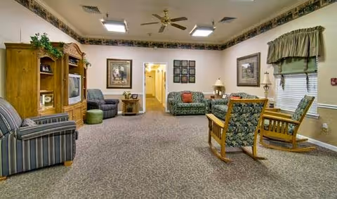 A cozy senior living facility common area with patterned carpet, multiple upholstered chairs and sofas, a wooden entertainment center with a TV, framed artwork on the walls, a ceiling fan, and a window with a valance.