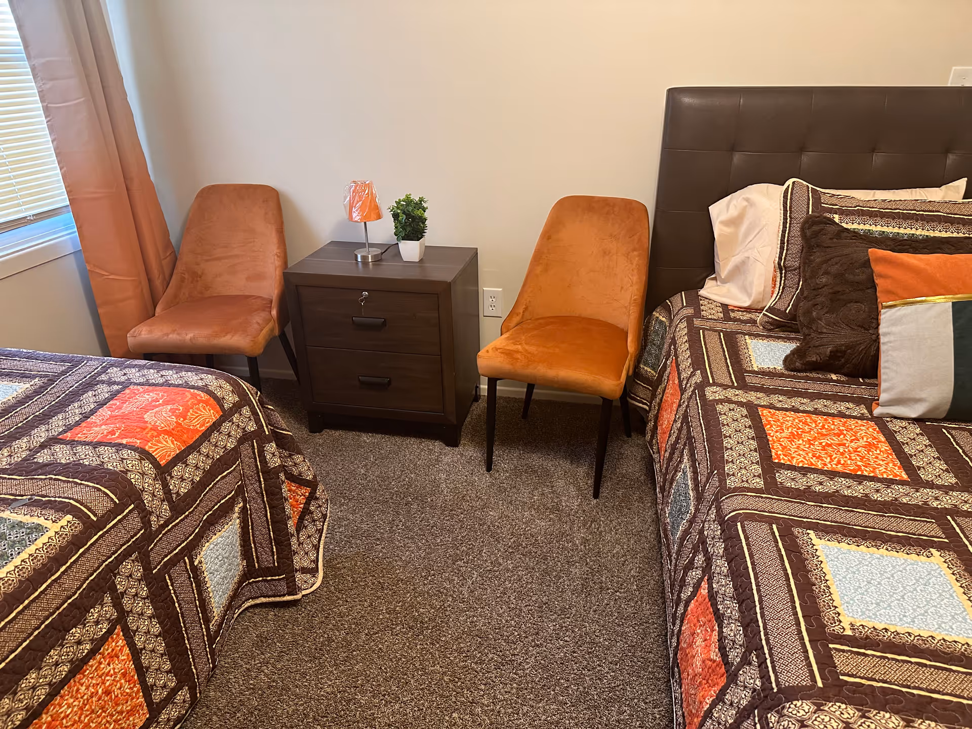 A bedroom with two beds covered in colorful patchwork quilts, two orange upholstered chairs, a dark wooden nightstand with two drawers, a small lamp with an orange shade, and a small green potted plant. There is a window with orange curtains on the left side.