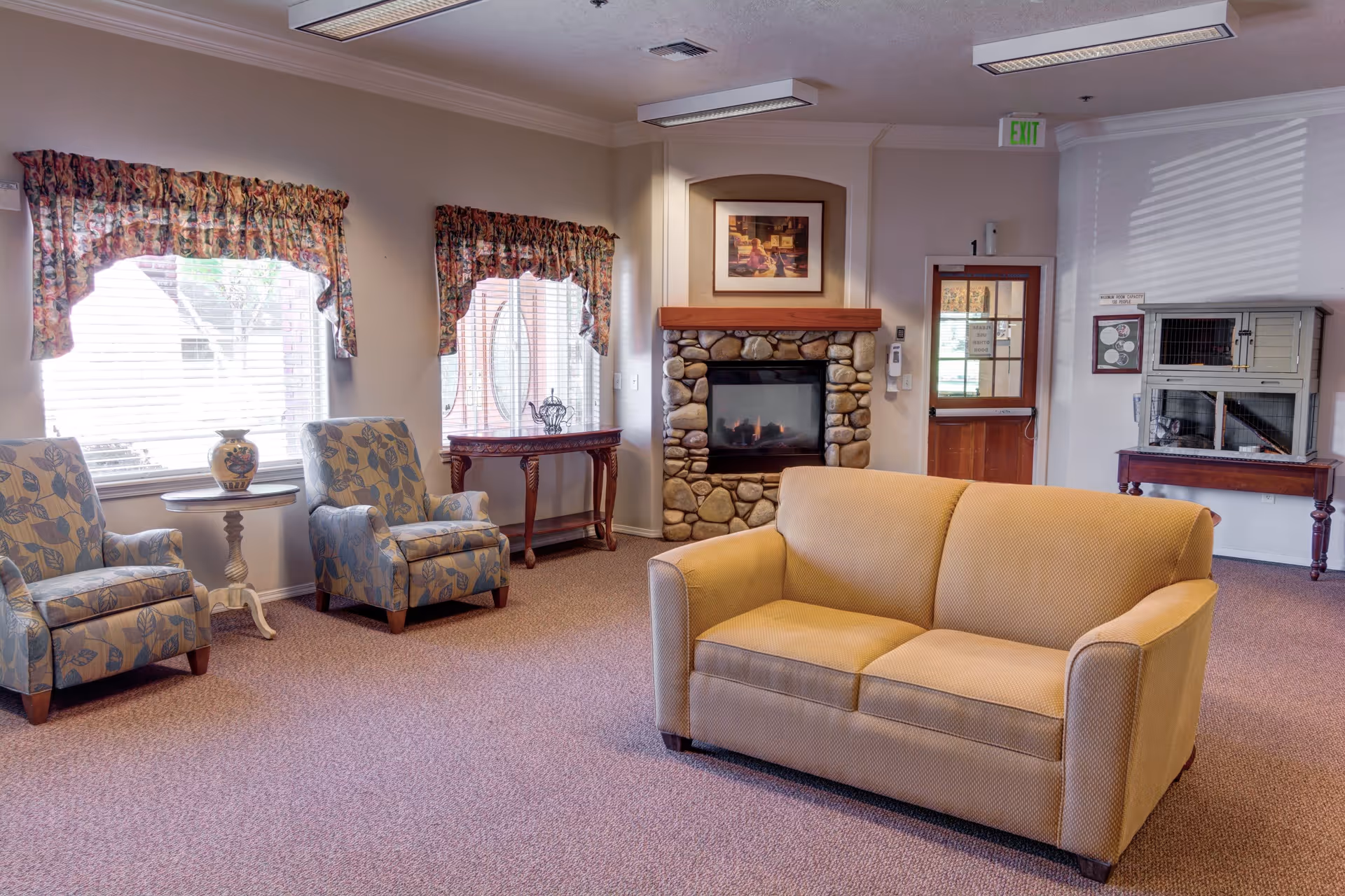 A cozy senior living facility common area with a beige couch in the center, two patterned armchairs near windows with floral valances, a stone fireplace with a framed picture above it, a wooden door, and a small table holding a pet cage on the right side.
