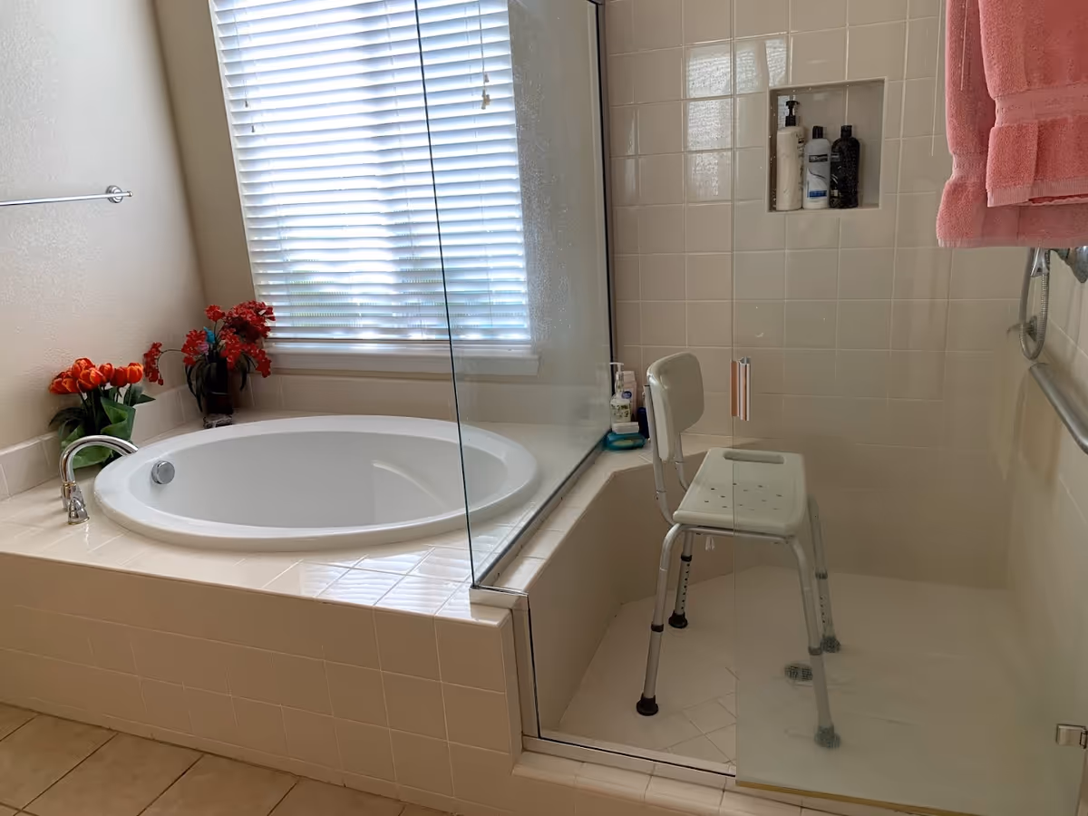 Tiled bathroom with a soaking tub beside a glass-enclosed shower containing a white shower chair and a pink towel.