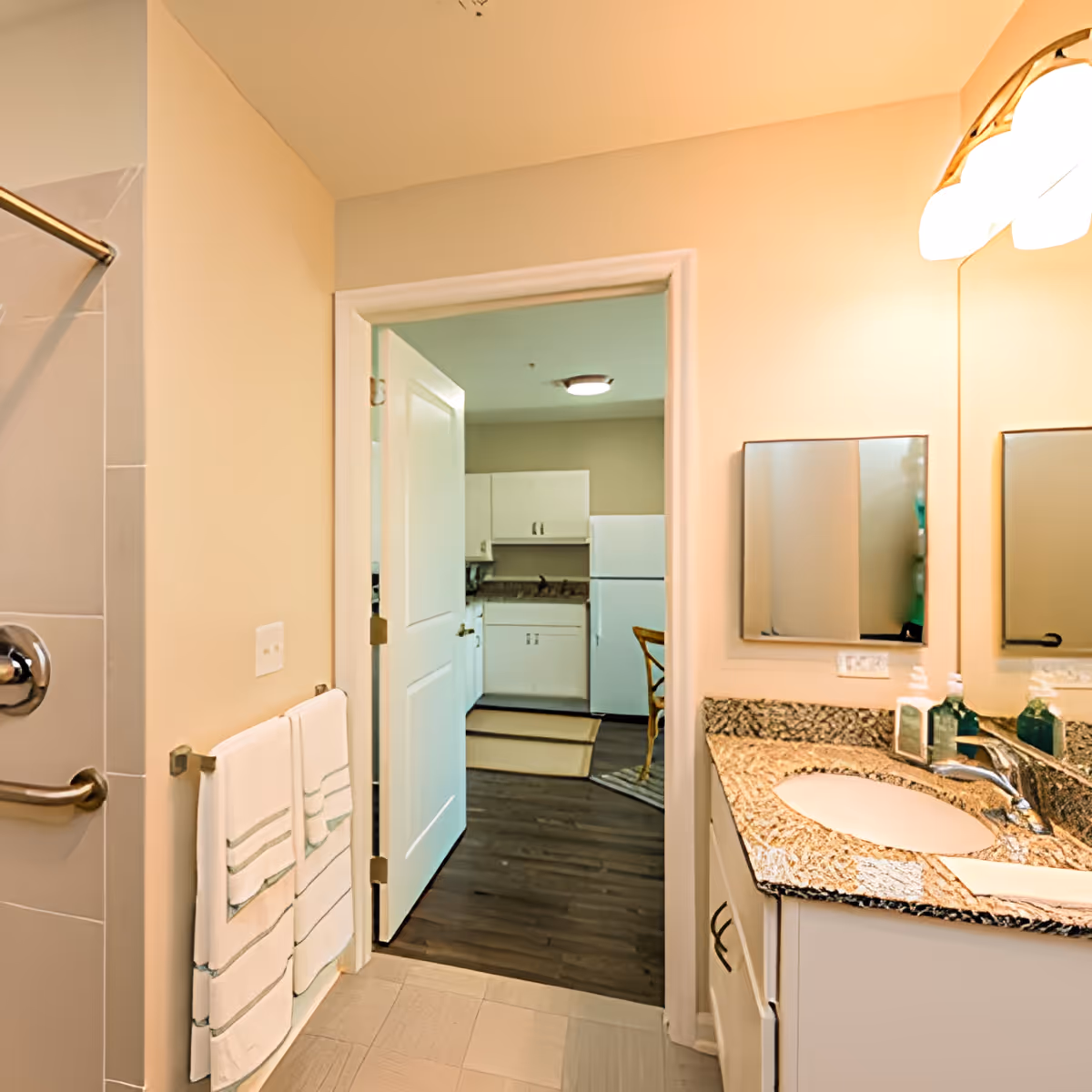 View of a bathroom with a granite countertop sink, mirror, and wall-mounted light fixture. Two white towels hang on a rack next to a shower with a grab bar. The bathroom door is open, showing a kitchen area with white cabinets, a refrigerator, and a wooden chair on dark wood flooring.