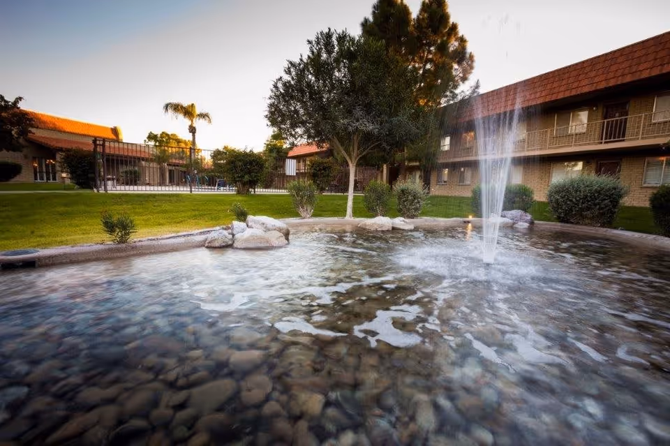 Outdoor view of a retirement facility featuring a small pond with a water fountain, surrounded by rocks and greenery. In the background, there are two-story buildings with balconies and a fenced swimming pool area, along with trees and well-maintained grass.